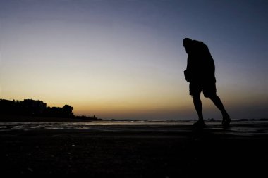 Gün batımında sahilde yürüyen bir adamın silüeti..
