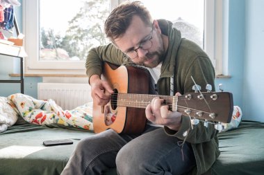 Akustik altı telli gitarlı konsantre bir adam evde yatakta otururken yeni bir melodi öğreniyor..