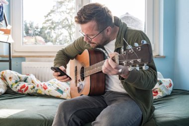 Akustik gitarlı bir adam evde bir yatakta otururken akıllı telefondaki notalara bakarken yeni bir melodi öğreniyor..