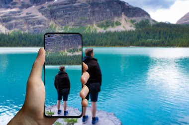 Tourist taking a picture with a mobile phone of a man standing at the lake Ohara, Canada.	