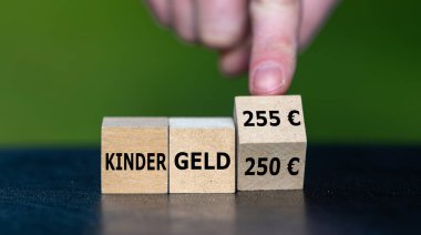 El, küpü döndürür ve Almanca 'Kindergeld 250 EUR' (çocuk nafakası 250 Euro) ifadesini 'Kindergeld 255 Euro' (çocuk nafakası 255 Euro) olarak değiştirir. Almanya 'da çocuk desteğindeki artışın sembolü.