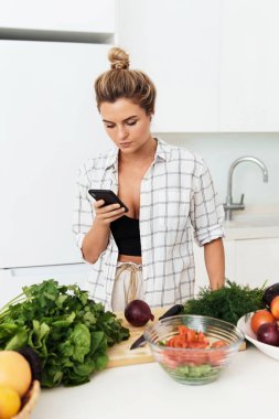 Kablosuz kulaklıklı genç ve güzel bir kadın modern beyaz mutfakta yemek pişirirken akıllı telefon kullanıyor.