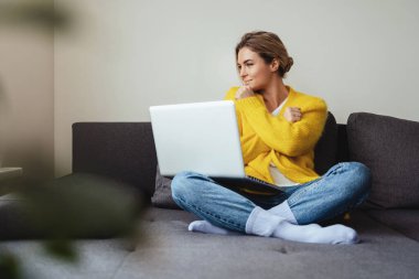 Sarı hırka giyen genç bir kadın kanepede oturuyor ve dizüstü bilgisayar kullanıyor.