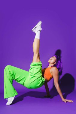 Dertsiz aktif kadın dansçı renkli spor giyerek mor arka plana karşı gösteri yapıyor.
