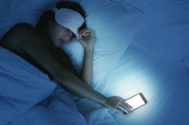 Uykulu kadın yatakta uzanıyor ve gece akıllı telefona bakıyor.