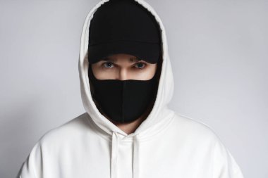 Beyaz kapüşonlu, siyah beysbol şapkalı ve gri arka planda kumaş maskeli genç bir adam.