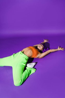Dertsiz aktif kadın dansçı renkli spor giyerek mor arka plana karşı gösteri yapıyor.