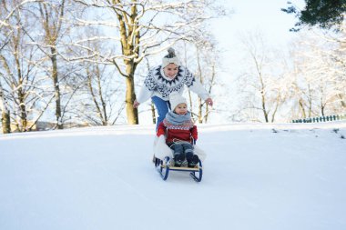 Annesi ve sıcak kazaklar giyen küçük tatlı oğlu güneşli kış gününde kızak tepesinde sevişiyorlar.