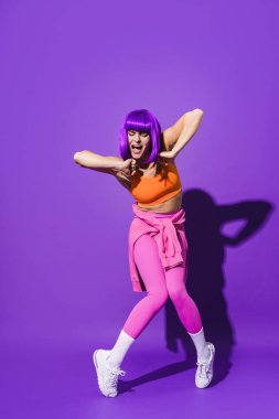 Dertsiz genç bayan dansçı renkli spor giyerek mor arka plana karşı gösteri yapıyor.