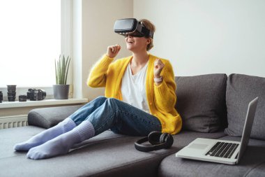 Kanepede oturup evde sanal gerçeklik kulaklığı takan heyecanlı genç bir kadın.