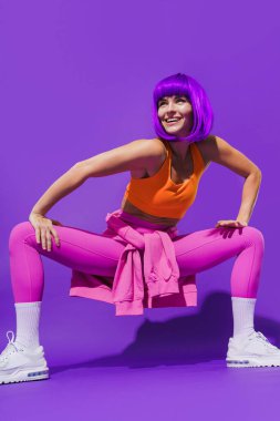 Dertsiz genç bayan dansçı renkli spor giyerek mor arka plana karşı gösteri yapıyor.