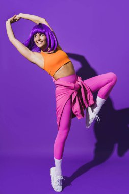 Dertsiz genç bayan dansçı renkli spor giyerek mor arka plana karşı gösteri yapıyor.