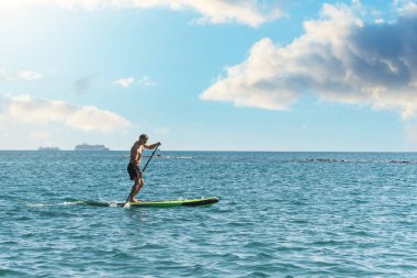 Genç erkek sörfçü ayakta kürek çekiyor ve okyanusta kürek çekiyor.