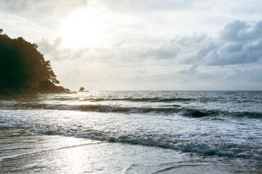 Köpüklü deniz dalgaları, yüksek gelgit sırasında kumlu bir plaja çarpıyor. Arka planda bulutlu bir gökyüzü var..