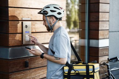 Sırtında yalıtımlı çanta olan genç ekspres kurye elinde telefon ve yakınlarda park edilmiş bisikletle dahili telefon kullanıyor..