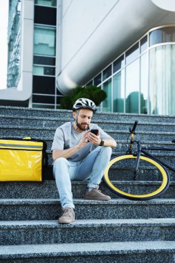 Genç ekspres yemek kuryesi izole edilmiş çanta ve bisikletle merdivenlerde oturuyor ve telefonuna bakıyor..