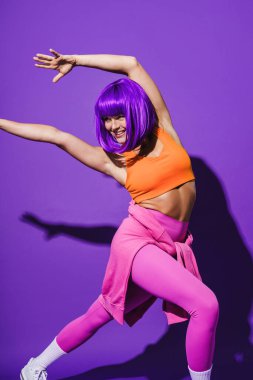 Dertsiz genç bayan dansçı renkli spor giyerek mor arka plana karşı gösteri yapıyor.