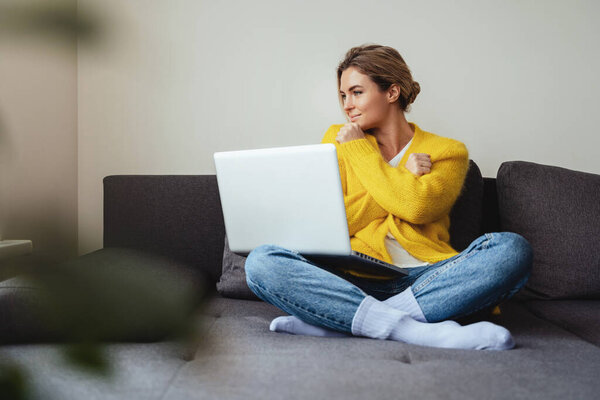 Young woman wearing yellow cardigan sitting on the sofa and using laptop computer