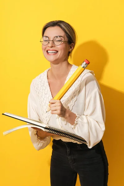 年轻而快乐的女人戴着眼镜 手里拿着大铅笔和笔记本 背景是黄色的 — 图库照片