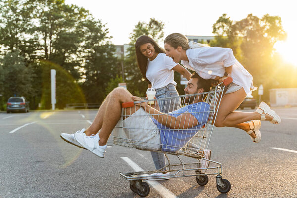 Трое друзей веселятся и катаются на тележке на парковке возле супермаркета в летний вечер
