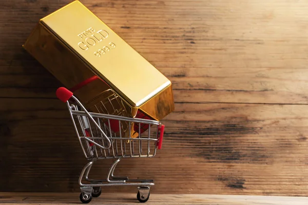 stock image Closeup shot of a large shiny gold bar put in a tiny shopping cart with wooden wall background.