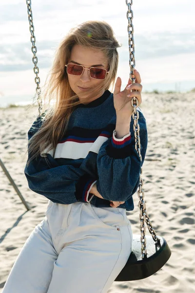 Stock image Young pretty woman is sitting on the swing and smiling at the sandy beach.