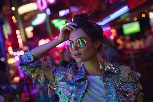 Young stylish woman wearing jacket with shining sequins on the city street with neon lights