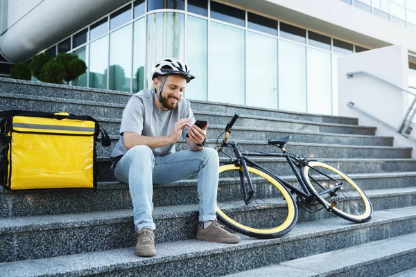 Young smiling express food delivery courier is sitting on the stairs with insulated bag and bicycle and looking at his phone.