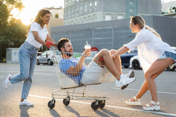 三个不同的朋友在夏天的傍晚在超级市场附近的停车场里玩乐和乘坐购物车 — 图库照片