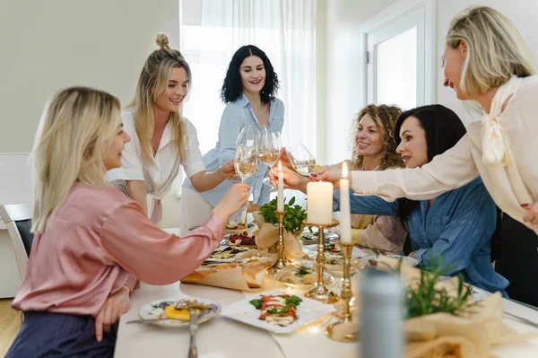 年轻而快乐的女性朋友们 在摆满食物和白葡萄酒 点缀着蜡烛的节日餐桌旁 碰碰着眼镜笑着 — 图库照片