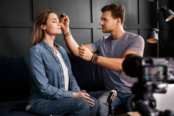 Молодой мужчина красоты блоггер сидит на диване перед камерой наносит макияж на красивую молодую женщину