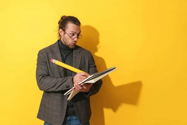 面白い髭の男を着て眼鏡を保持大きな鉛筆とノートブック上の黄色の背景 — ストック写真