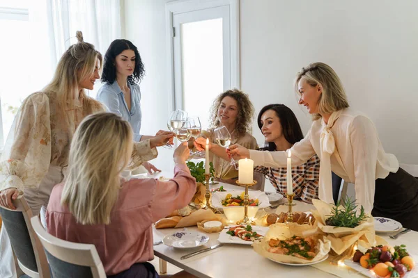 年轻快乐的女性朋友们在摆满食物和白葡萄酒 点缀着蜡烛的节日餐桌前 碰杯微笑着 — 图库照片