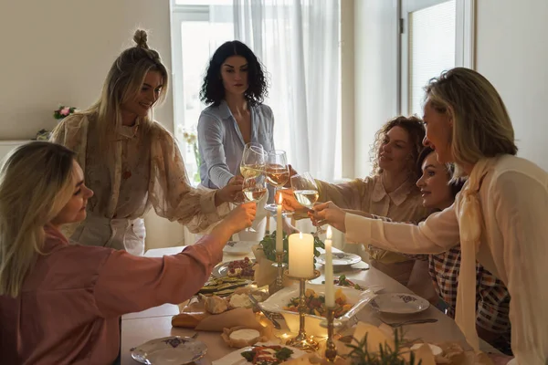 年轻快乐的女性朋友们在摆满食物和白葡萄酒 点缀着蜡烛的节日餐桌前 碰杯微笑着 — 图库照片