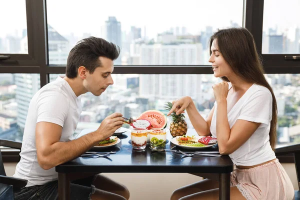 Young happy couple eating healthy breakfast in modern apartment with large windows and city view