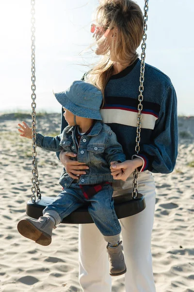若いですお母さんと彼女の小さな息子座っているスイング離れて見ている砂のビーチで — ストック写真