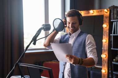 Genç, sakallı yakışıklı bir adam, arkasında makyaj aynası olan bir piyanonun önündeki müzik stüdyosunda gülümsüyor..