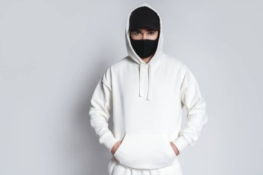 Beyaz kapüşonlu, siyah beysbol şapkalı ve gri arka planda kumaş maskeli genç bir adam.