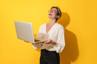 Gözlüklü neşeli genç bayan sarı arka planda dizüstü bilgisayar kullanıyor.