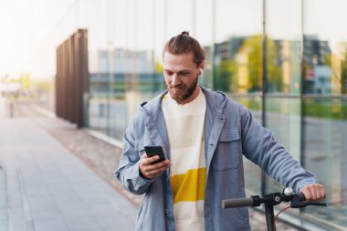 Akıllı telefonunda scooter sistemi uygulaması kullanan, e-scooter kullanan genç bir adam.