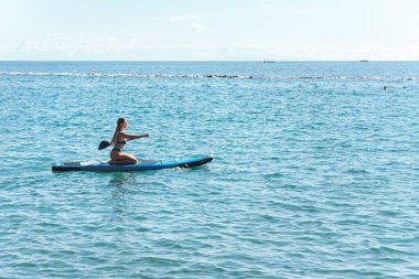 Bikinili genç seksi sörfçü bir raket sürüyor ve okyanusta kürek çekiyor..