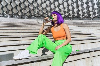 Kaygısız genç kadın renkli spor kıyafetleri giyerek yaz sporundan sonra su içiyor.