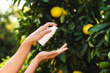 Kadının elinde beyaz bir şişe cilt bakımı ürünü var. Bulanık limon ağacı arka planında..