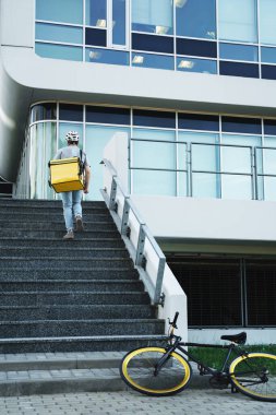 Arkasında yalıtılmış çanta taşıyan genç ekspres yemek kuryesi bisikletini park ettikten sonra merdivenlerden çıkıyor..