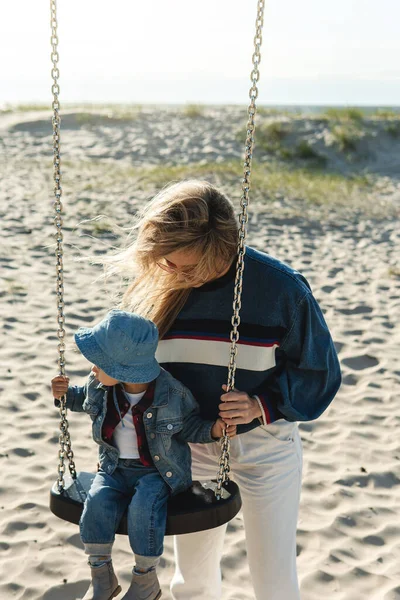 若いです笑顔母あります持っています彼女の小さな息子座っています上のスイングでザ砂のビーチ — ストック写真