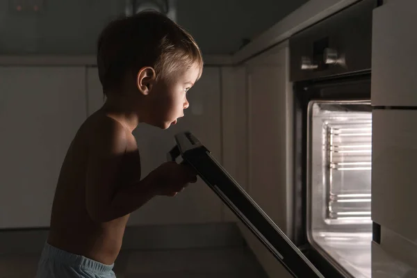 Cute curious baby boy opens the hot oven. Concept of safety and possible problems with unattended children.