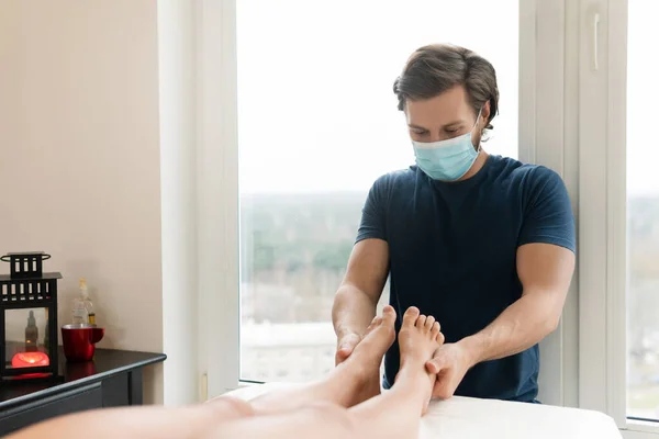 Professional man masseur wearing prevention mask during female foot massage session