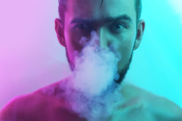 Portrait of handsome young man with wet skin in ultraviolet light smoking vape