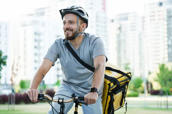 Young smiling express food delivery courier is riding a bicycle with an insulated bag behind his back.