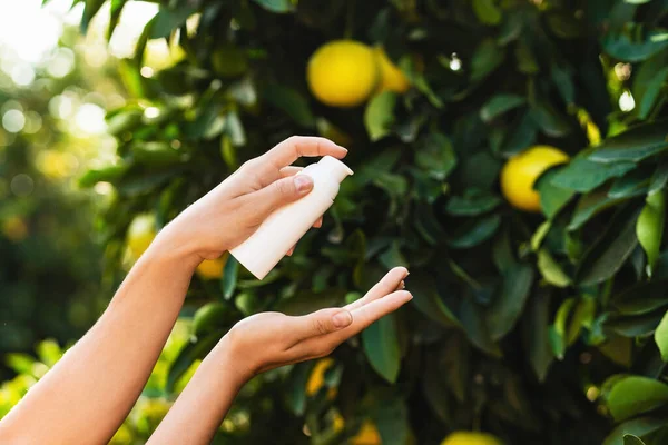 stock image Woman holds a white bottle of skin care product in her hands on blurry lemon tree background.